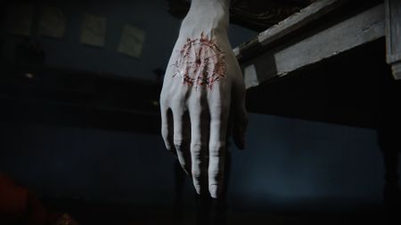 The Duskbloods screenshot on she Nintendo Switch 2, showing a limp hand from someone who appears to be dead, with a strange symbol carved into the hand